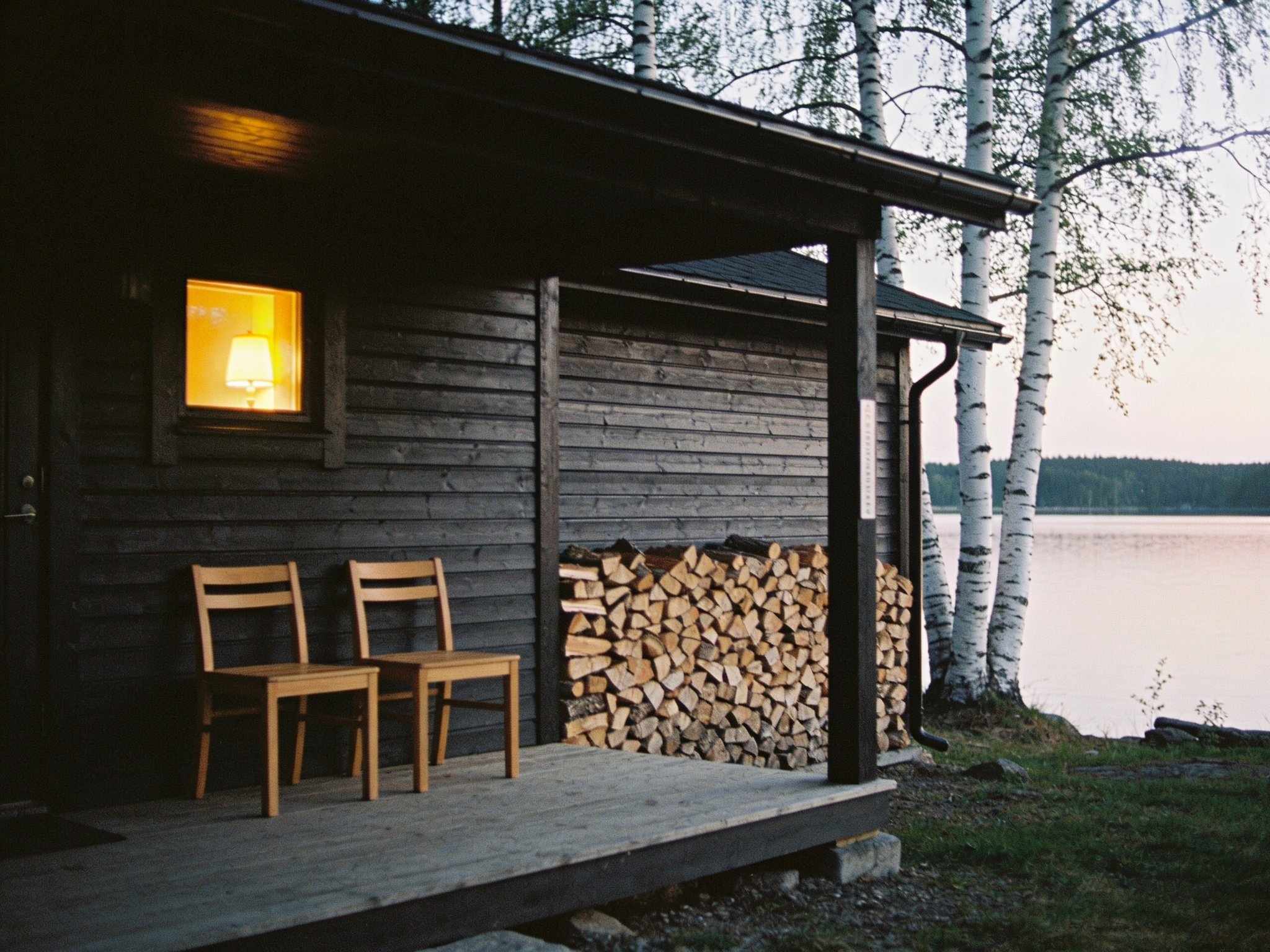 A dark timber cabin with two wooden chairs and a stack of firewood on its porch, a lamp glowing in the window, birches and the lake beside it at dusk