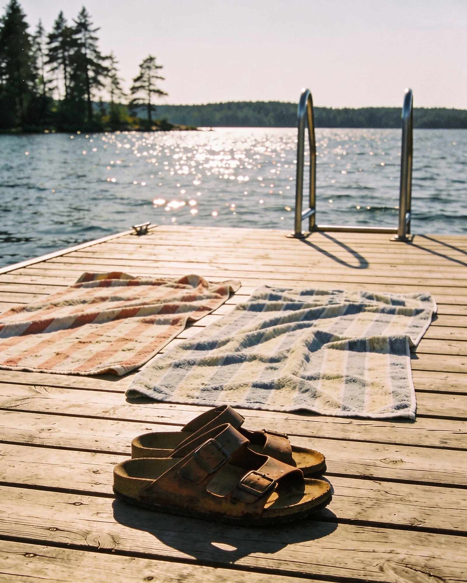Two striped towels and a pair of worn leather sandals on a sunlit wooden jetty, with a swim ladder and the sparkling lake beyond