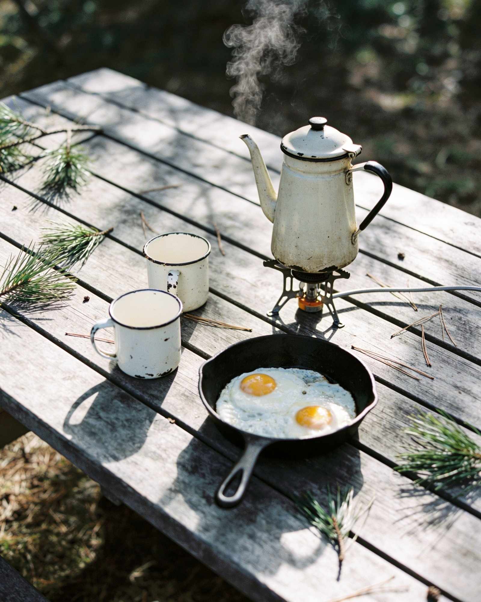 An enamel coffee pot steaming on a small camping stove beside two chipped enamel mugs and a cast-iron pan of fried eggs on a weathered picnic table